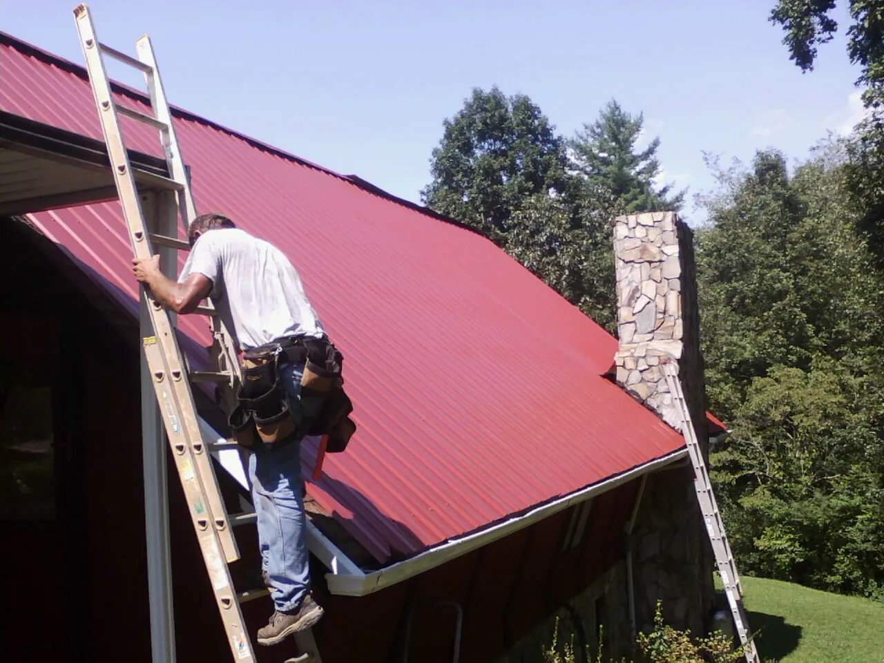 Roofer on a ladder beside a red metal roof and stone chimney — Soffit & Fascia Repair by Roofing Brothers in Prattsburgh